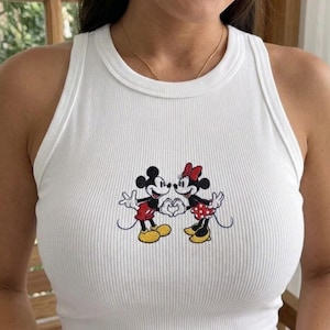 Può includere: Canotta bianca a coste con un'immagine ricamata di Topolino e Minnie Mouse. Topolino indossa pantaloncini rossi e Minnie indossa una gonna a pois rossi. I personaggi dei cartoni animati formano un cuore con le mani.