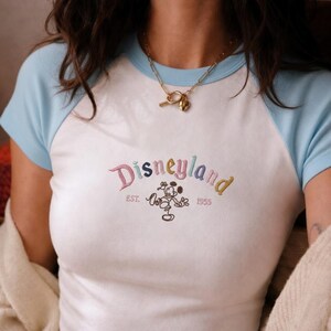 Puede incluir: Camiseta ragl&aacute;n blanca y azul claro con la palabra "Disneyland" bordada en hilo colorido. La camiseta presenta un gr&aacute;fico de Mickey Mouse y el texto "EST. 1955". Se lleva un collar dorado alrededor del cuello.