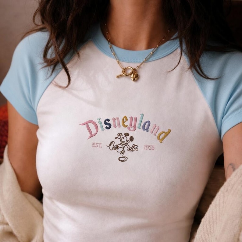 Puede incluir: Camiseta ragl&aacute;n blanca y azul claro con la palabra "Disneyland" bordada en hilo colorido. La camiseta presenta un gr&aacute;fico de Mickey Mouse y el texto "EST. 1955". Se lleva un collar dorado alrededor del cuello.