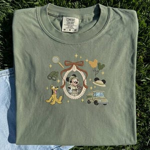 May include: Olive green Comfort Colors t-shirt with an embroidered design of Mickey Mouse in safari attire, Pluto, binoculars, a magnifying glass, and a safari vehicle. The shirt has a crew neck and short sleeves.
