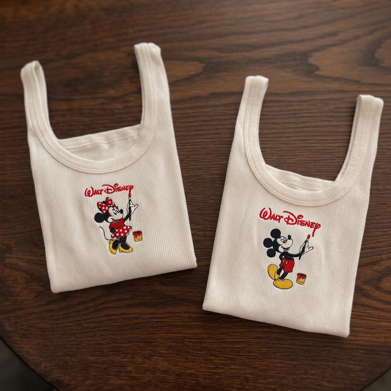 May include: Two off-white tank tops with the Walt Disney logo and a cartoon character design. One features Minnie Mouse in a red polka-dot dress, and the other features Mickey Mouse in red shorts. Both are holding paintbrushes.
