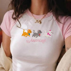 May include: A white and light pink raglan t-shirt featuring an embroidered graphic of three kittens from The Aristocats. The design includes an orange kitten, a gray kitten, and a white kitten with a pink bow, with the text The Aristocats below.