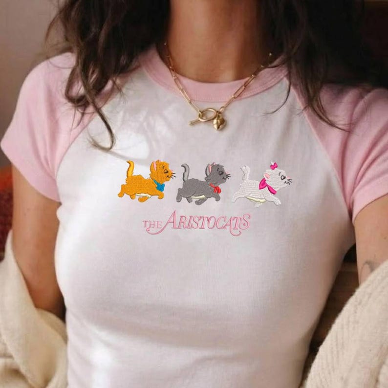 May include: A white and light pink raglan t-shirt featuring an embroidered graphic of three kittens from The Aristocats. The design includes an orange kitten, a gray kitten, and a white kitten with a pink bow, with the text The Aristocats below.
