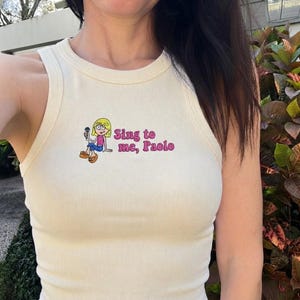May include: A cream-colored ribbed tank top featuring an embroidered graphic of a blonde character holding a microphone, accompanied by the pink embroidered text "Sing to me, Paolo" positioned to the right of the character illustration.