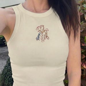K&ouml;nnte beinhalten: Cremefarbenes geripptes Tanktop mit einer Cartoon-Pferde-Stickerei. Das Pferd tr&auml;gt einen Cowboyhut und hat ein blau-wei&szlig; geflecktes Accessoire. Das Top ist &auml;rmellos und hat einen runden Ausschnitt.