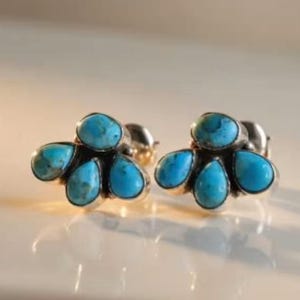 May include: A pair of silver stud earrings featuring a cluster of five teardrop-shaped, turquoise-colored stones. The earrings are displayed on a light-colored surface, showcasing their bright blue color and intricate design. The earrings are a jewelry item.