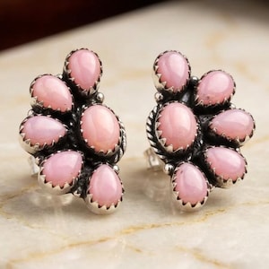 May include: A pair of stud earrings featuring multiple teardrop-shaped pink stones arranged in a fan-like cluster around a central oval pink stone. The stones are set in silver metal with a serrated edge detail around each individual setting.