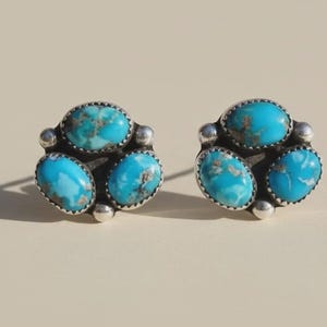May include: A pair of silver stud earrings, each featuring three oval turquoise stones. The bright blue stones have brown matrix patterns and are set in a silver bezel. The earrings are displayed on a neutral background.