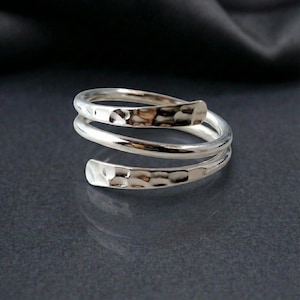 May include: A silver-colored, spiral ring with a hammered texture. The ring wraps around the finger twice, with a flat, textured end on each side. The ring is displayed on a dark surface, highlighting its metallic shine.