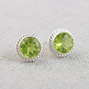 May include: A pair of round stud earrings featuring a vibrant green gemstone. The gemstones are set in a silver-colored metal with a twisted rope design around the perimeter. The earrings are displayed on a light gray surface.