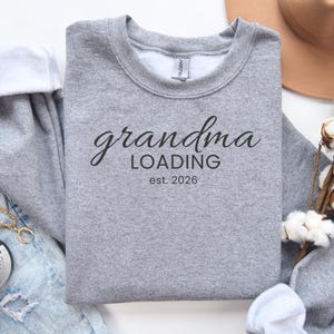 May include: A heather gray sweatshirt with the words "grandma LOADING est. 2026" printed in black script. The sweatshirt has a crew neck and long sleeves. The text indicates an upcoming event.