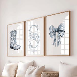 May include: Three framed wall art prints featuring a blue and white floral pattern. The left print shows a cowboy boot, the centre print displays the letter S with the name Sophia written below, and the right print depicts a large decorative bow.