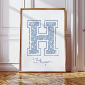 May include: A framed print featuring a large, light blue letter "H" filled with a floral pattern. The name "Harper" is written in cursive below the letter. The frame is a light brown color.