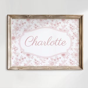 May include: A framed print with the name "Charlotte" in pink script. The background is a floral design in pink and white, set within a decorative, light pink border. The frame is made of light-colored wood, creating a vintage feel.