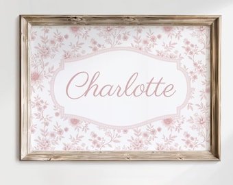 May include: A framed print featuring the name "Charlotte" in a soft pink script. The background is a delicate floral pattern in pink and white, framed by a rustic wooden border. The overall design is elegant and feminine.
