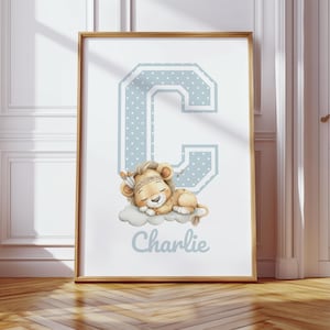 Puede incluir: Un estampado enmarcado con la letra "C" azul claro con lunares blancos, y un cachorro de león durmiendo con un tocado de plumas. El nombre "Charlie" está escrito debajo en azul claro. La obra de arte está en un marco de madera.