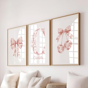 Op de afbeelding: Drie ingelijste prints met een lichte houten afwerking. De prints tonen aquarel illustraties van een strik, de letter "C" met bloemmotief en kersen, allemaal in roze en rode tinten. De letter "C" heeft de naam "Charlotte" eronder.