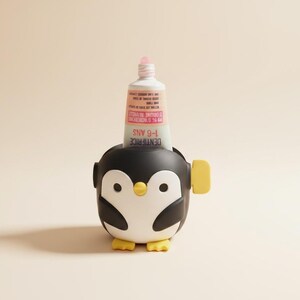 Puede incluir: Un exprimidor de tubos de pasta de dientes con forma de pingüino en blanco y negro, con pies amarillos y una perilla lateral amarilla. Un tubo de pasta de dientes blanco con texto rosa y negro que dice DENTIFRICE 1-6 ANS insertado.