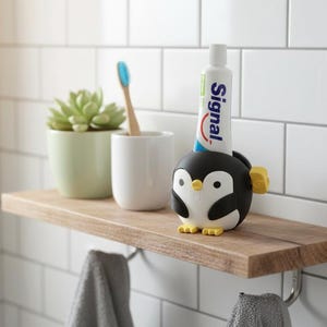 Op de afbeelding: Een houten plank in een badkameromgeving bevat een tandenborstel, een kleine succulent in een pot en een witte beker. Een zwart-witte tandpasta-houder in de vorm van een pinguïn met een tube Signal-tandpasta staat ook op de plank.