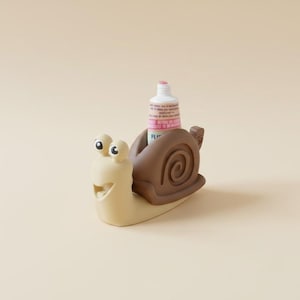 Puede incluir: Un exprimidor de pasta de dientes con forma de caracol beige y concha marrón oscuro. El caracol tiene dos ojos en tallos y una boca abierta. Un tubo de pasta de dientes blanco con texto rosa y azul está insertado en la concha marrón.