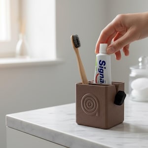 Puede incluir: Un portacepillos marrón con un cepillo de dientes de bambú y un tubo de pasta de dientes. El tubo de pasta de dientes tiene la marca "Signa" impresa. Una mano alcanza el tubo de pasta de dientes.