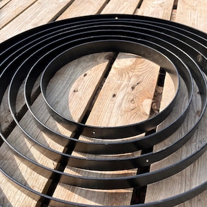 May include: A collection of black metal rings of varying sizes, arranged in a circular pattern. The rings are set against a backdrop of weathered wooden planks, creating a contrast in textures and tones. The image is well-lit, highlighting the smooth surfaces of the rings.