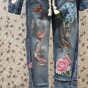 Può includere: Jeans in denim blu con varie toppe di tessuto e decorazioni, tra cui fiori, farfalle e bordini in pizzo.