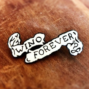 May include: A white wooden pin with the words "Wino Forever" written on it in black lettering. The pin is shaped like a banner.
