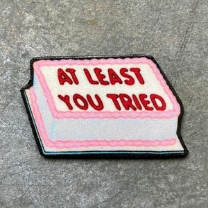 May include: A pink and white cake with black frosting and the text "At Least You Tried" in red.