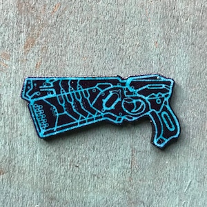 May include: A black and blue enamel pin depicting a futuristic gun with a circuit board design.
