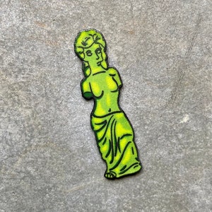Könnte beinhalten: Eine grüne und gelbe Statue einer Frau mit einem drapierten Gewand. Die Statue steht in einer stehenden Pose mit einem erhobenen Arm.