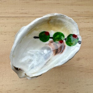 May include: A white seashell with a painted design of green olives with red centers on a black stem. The shell is on a light brown wooden surface.