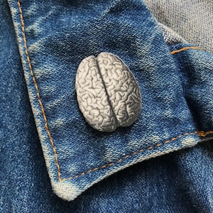 Könnte beinhalten: Eine weiße und graue Gehirn-förmige Anstecknadel auf einer blauen Jeansjacke.