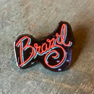 Op de afbeelding: Een zwarte emaille pin met het woord "Brazilië" geschreven in een gestileerd lettertype met rode en blauwe neon contouren.