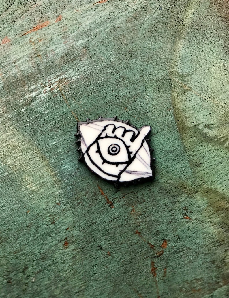 May include: A white enamel pin with a black outline depicting an eye with a hand reaching out from it. The eye is surrounded by a circle of spikes.