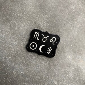 May include: Black and white zodiac sign charm with a black background and white symbols. The symbols include the zodiac signs for Scorpio, Taurus, Leo, a sun, a crescent moon, and a pine tree.