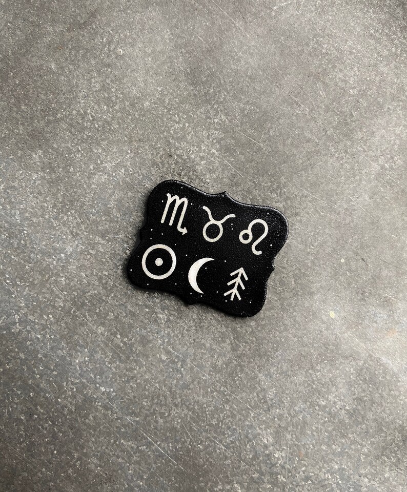 May include: Black and white zodiac sign charm with a black background and white symbols. The symbols include the zodiac signs for Scorpio, Taurus, Leo, a sun, a crescent moon, and a pine tree.