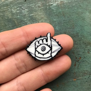 May include: White enamel pin with a black outline of an eye with a hand making a peace sign in front of it. The eye has a black pupil and a white iris.