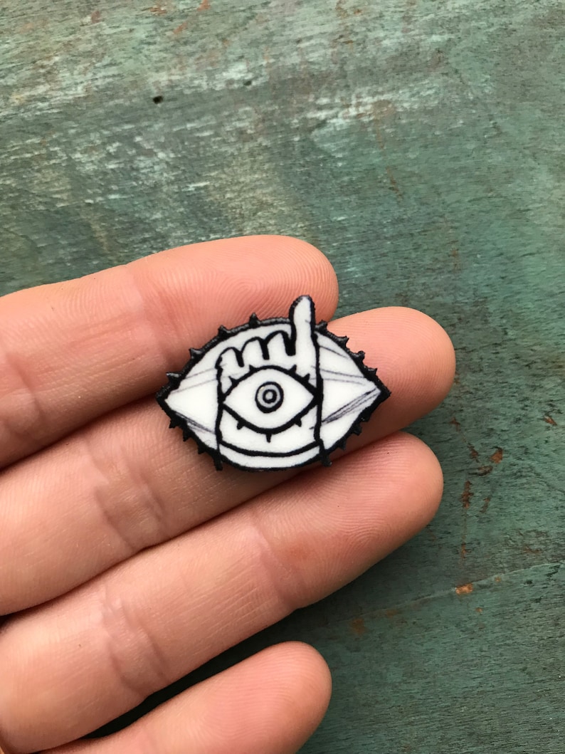May include: White enamel pin with a black outline of an eye with a hand making a peace sign in front of it. The eye has a black pupil and a white iris.