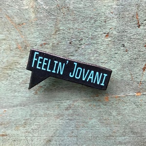Puede incluir: Pin de burbuja de di&aacute;logo en blanco y negro con el texto "Feelin' Jovani" en azul.