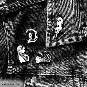 Puede incluir: Cuatro pines de esmalte blanco y negro en una chaqueta vaquera. Los pines representan a una mujer con una luna creciente, una mujer con el pelo largo, un hombre con cara de gato y un gato con una calavera.