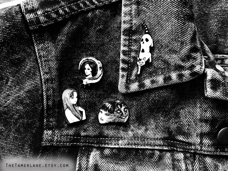 Puede incluir: Cuatro pines de esmalte blanco y negro en una chaqueta vaquera. Los pines representan a una mujer con una luna creciente, una mujer con el pelo largo, un hombre con cara de gato y un gato con una calavera.