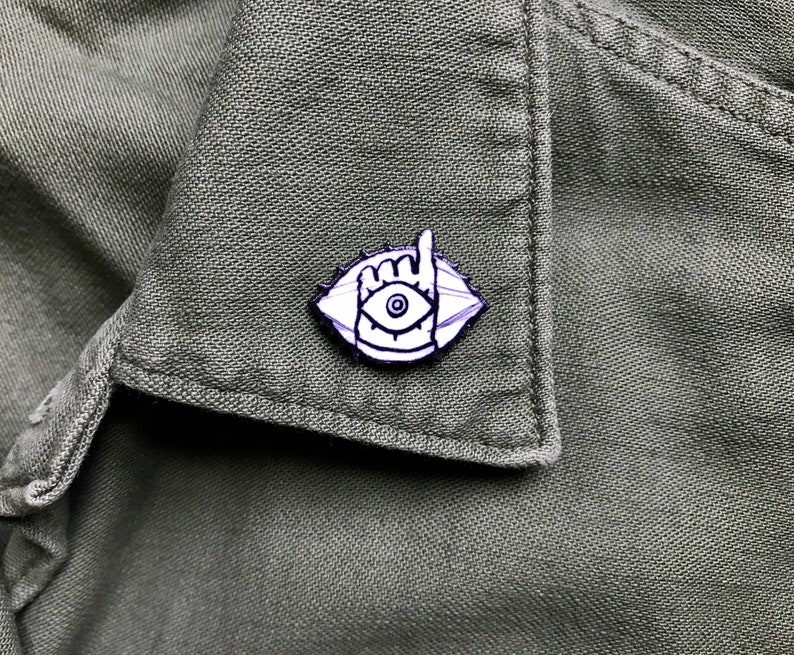 May include: White enamel pin with a black outline of an eye with a hand giving the 'rock on' sign. The eye is surrounded by a circle with a black outline.