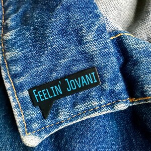 Puede incluir: Pin de esmalte negro y azul con el texto "Feelin' Jovani" en forma de bocadillo.