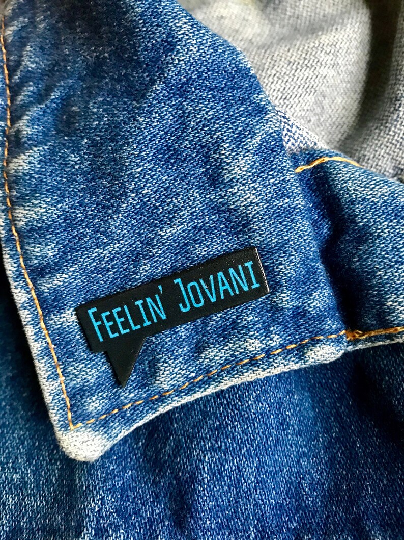 Puede incluir: Pin de esmalte negro y azul con el texto "Feelin' Jovani" en forma de bocadillo.