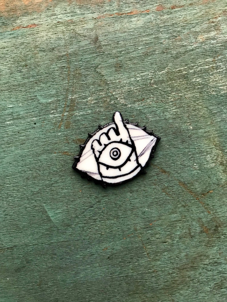 May include: White enamel pin with a black outline of an eye with a hand in front of it. The hand is making a shaka sign.