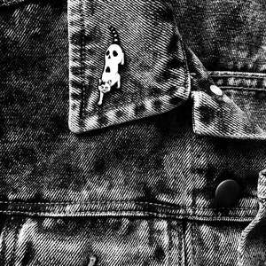 Puede incluir: Pin de esmalte blanco y negro de un gato con una expresi&oacute;n espeluznante. El pin est&aacute; sujeto a una chaqueta vaquera.