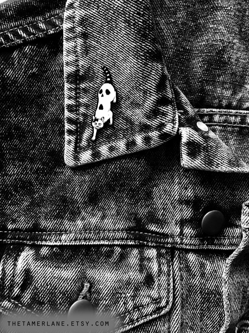 Puede incluir: Pin de esmalte blanco y negro de un gato con una expresi&oacute;n espeluznante. El pin est&aacute; sujeto a una chaqueta vaquera.