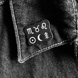 May include: Black and white enamel pin with astrological symbols including the zodiac signs of Scorpio, Taurus, and Leo, a crescent moon, a star, and a pine tree.