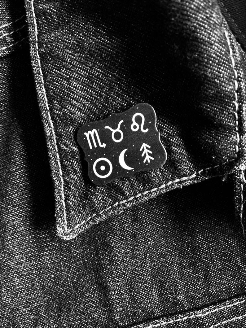 May include: Black and white enamel pin with astrological symbols including the zodiac signs of Scorpio, Taurus, and Leo, a crescent moon, a star, and a pine tree.